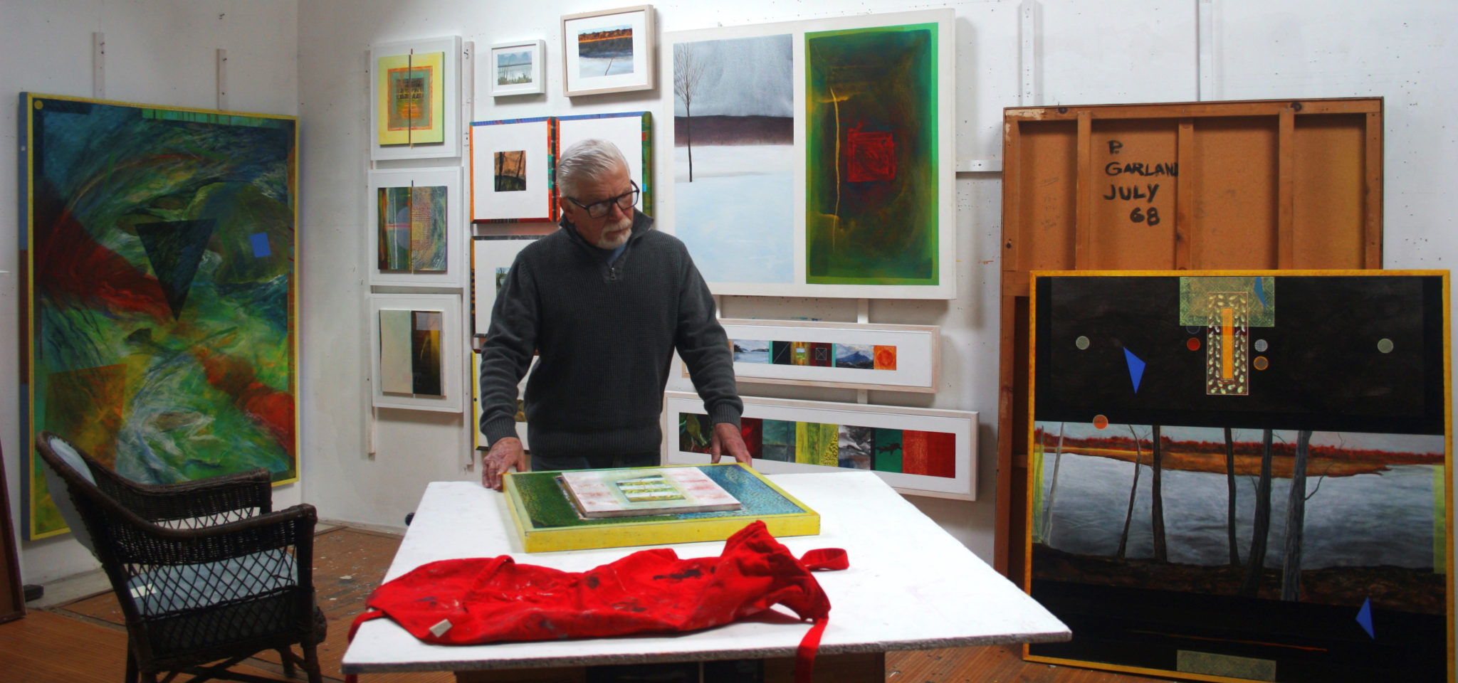 Paul Garland standing at a table, looking down at one of his paintings, in the middle of his studio.