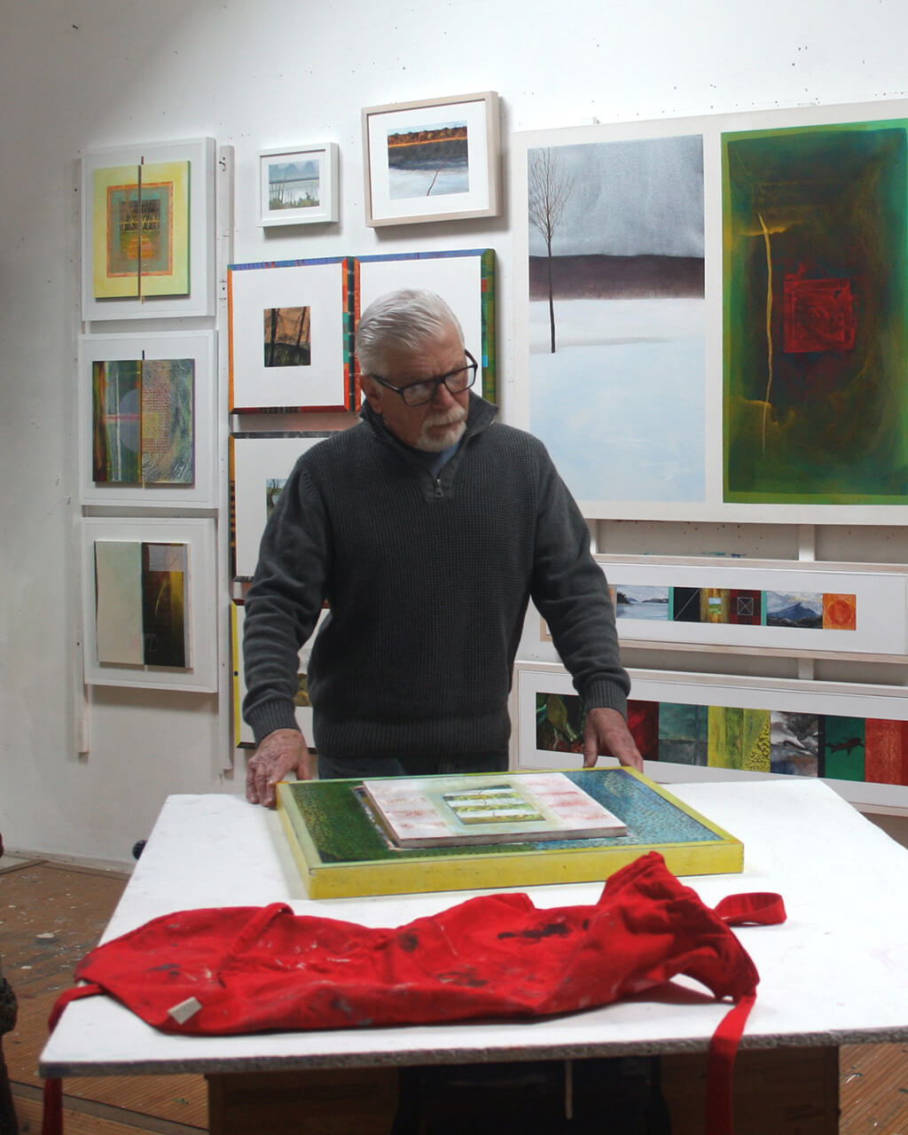 Paul Garland standing at a table, looking down at one of his paintings, in the middle of his studio.