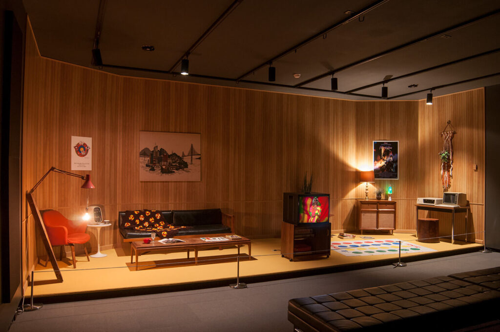 An installation of a mid-century American living room with furniture and a TV, within a darkened gallery space.