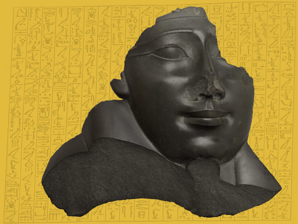 A broken sculpture of a face from a sarcophagus, made of black stone, against a yellow background covered in hieroglyphs.