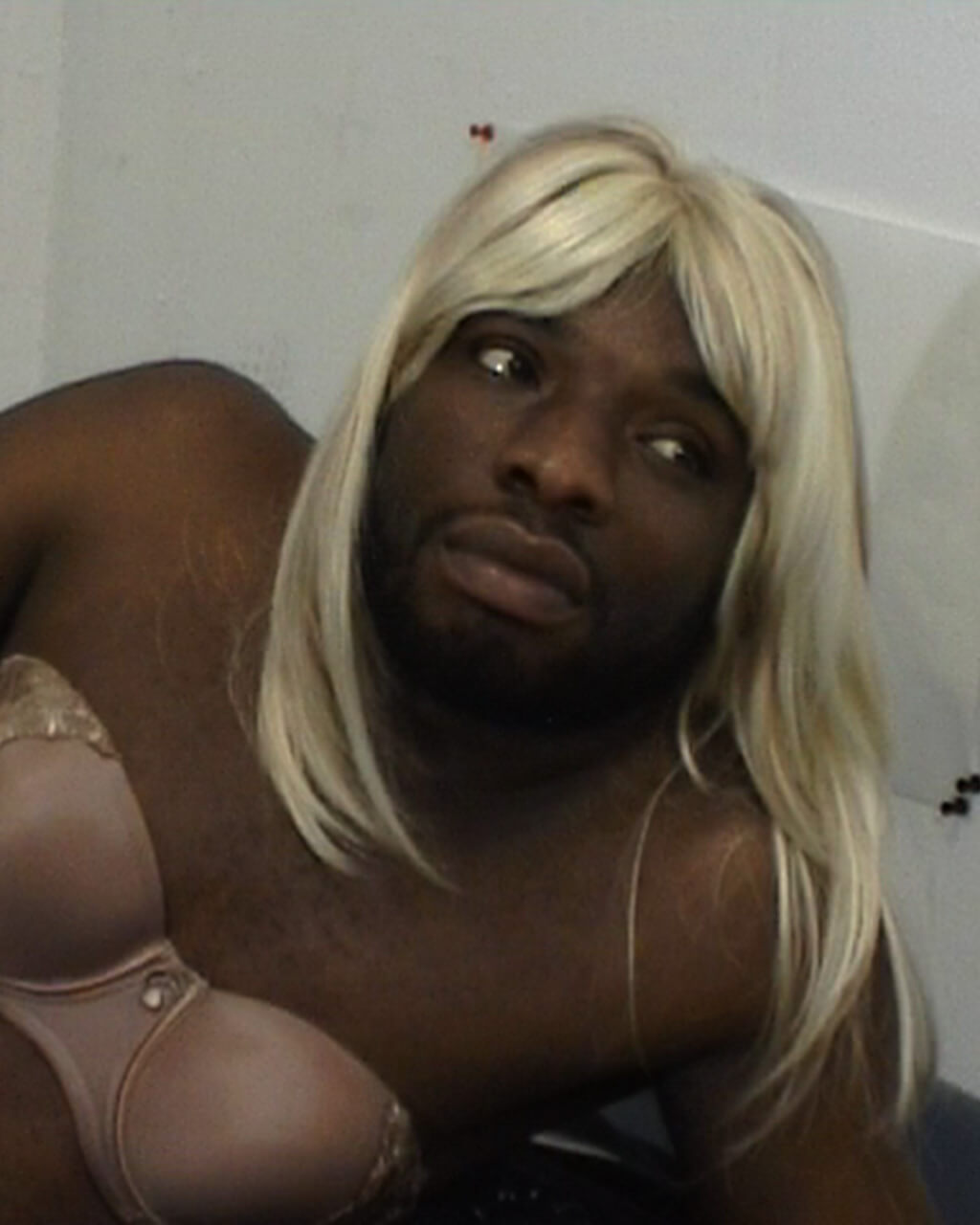 Kalup Linzy crossdressing as his alter ego, Katonya, wearing a blonde wig and a strapless bra, sprawled on the ground.