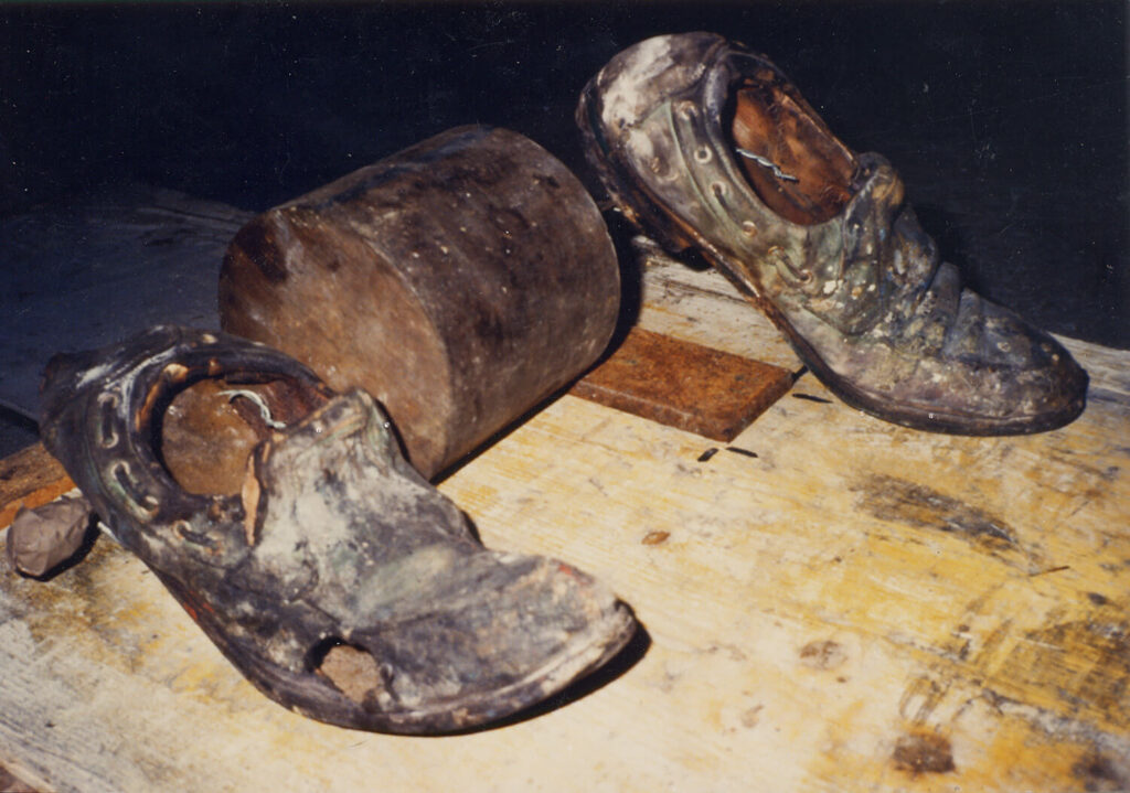 A contraption created from a dirty pair of shoes attached to a heavy cylinder on a wooden board.