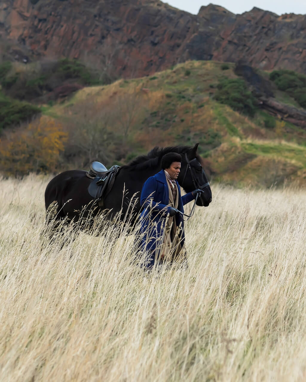 A black man walks a black horse across a field, mountains in the background.