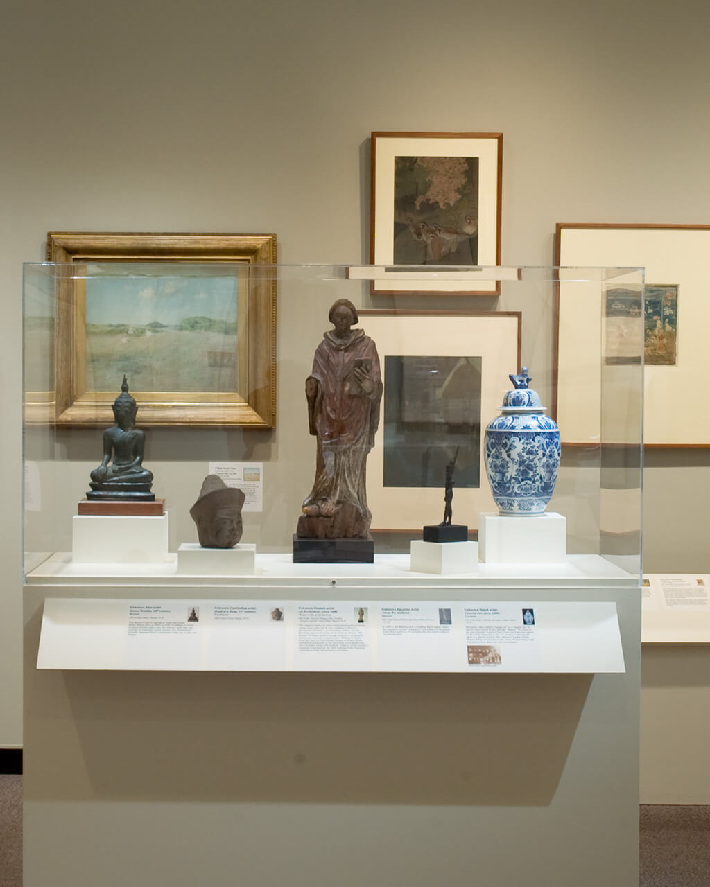 Framed works of art in multiple mediums on a gallery wall, with various small sculptures in a display case in front of the wall.