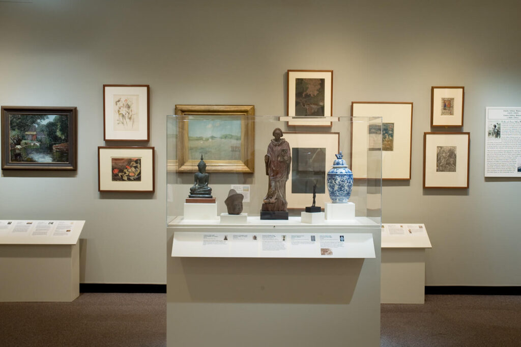 Framed works of art in multiple mediums on a gallery wall, with various small sculptures in a display case in front of the wall.