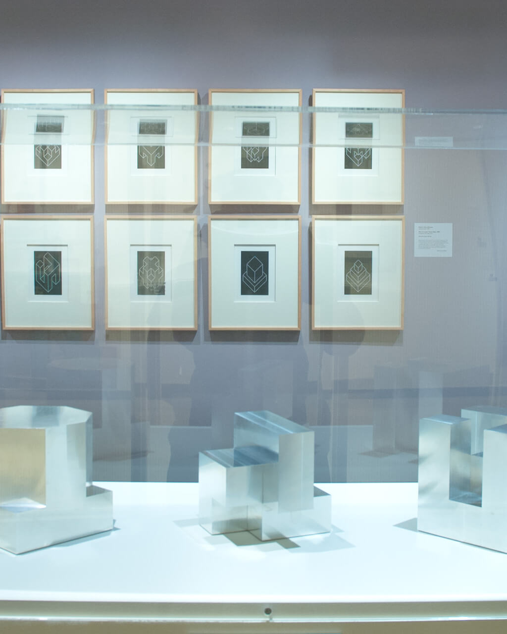 Small, framed prints hang on a lilac-painted wall in a gallery. In front of them stands a case with three silver geometric forms in it.