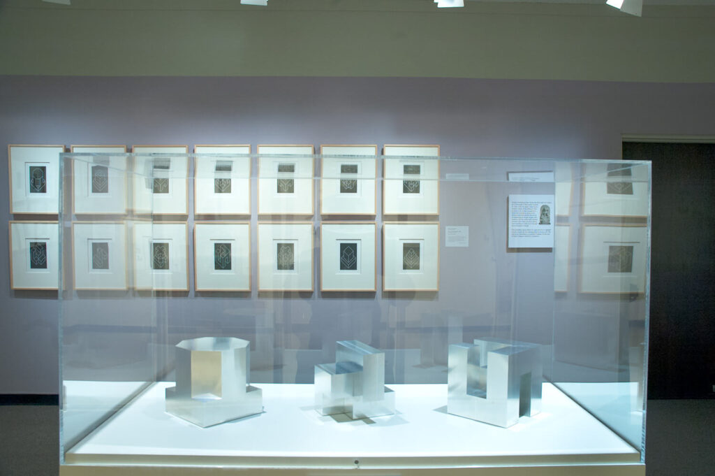 Small, framed prints hang on a lilac-painted wall in a gallery. In front of them stands a case with three silver geometric forms in it.