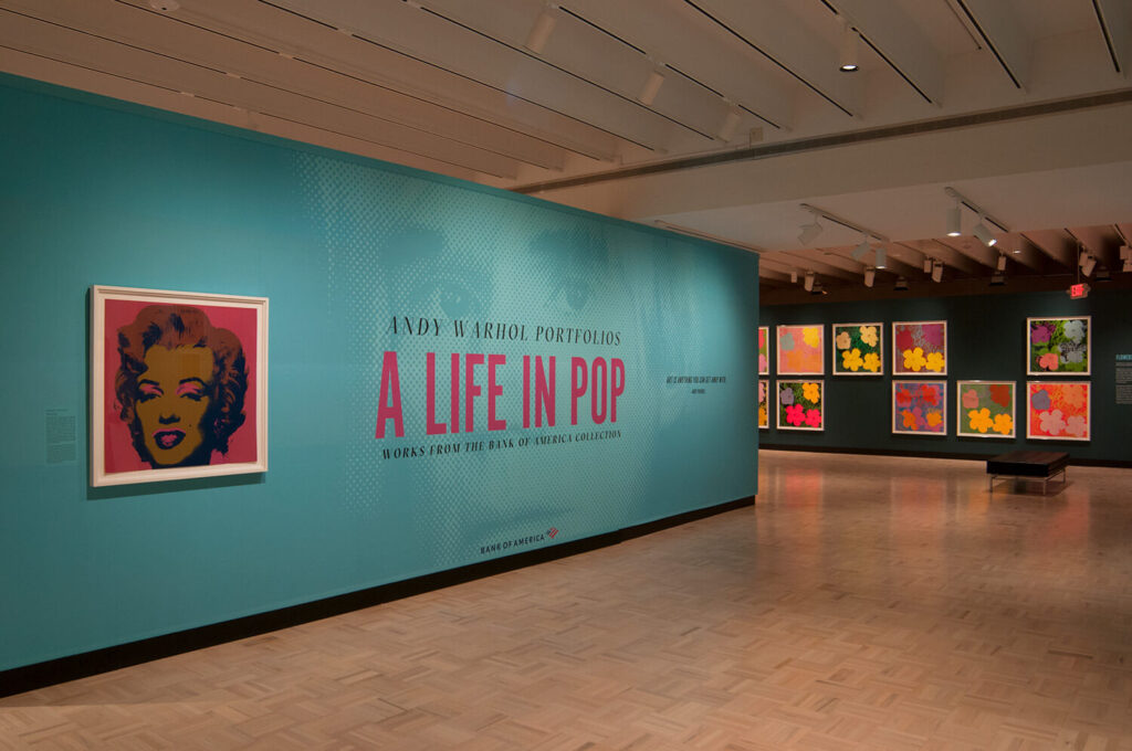 The title wall of the exhibition "Andy Warhol Portfolios: A Life in Pop," with some prints on a wall in the background.
