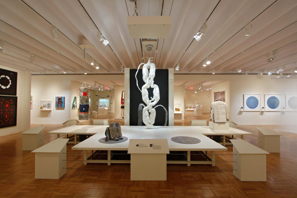 Art in many different mediums hanging on walls, the ceiling, or resting on a large platform in the middle of a white-walled gallery.