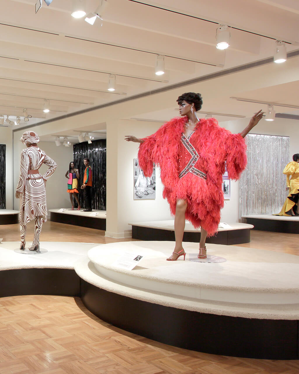 Gowns on mannequins positioned on a long, catwalk-like display platform in a large gallery.