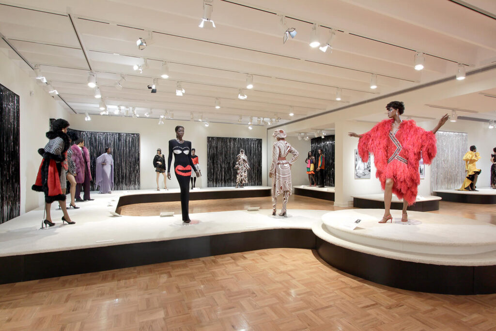 Gowns on mannequins positioned on a long, catwalk-like display platform in a large gallery.