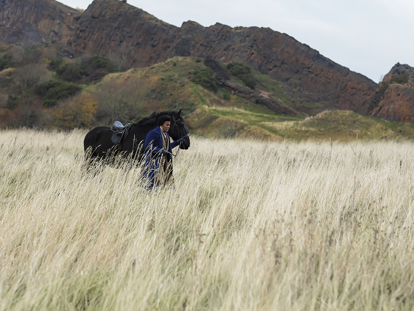 A black man walks a black horse across a field, mountains in the background.