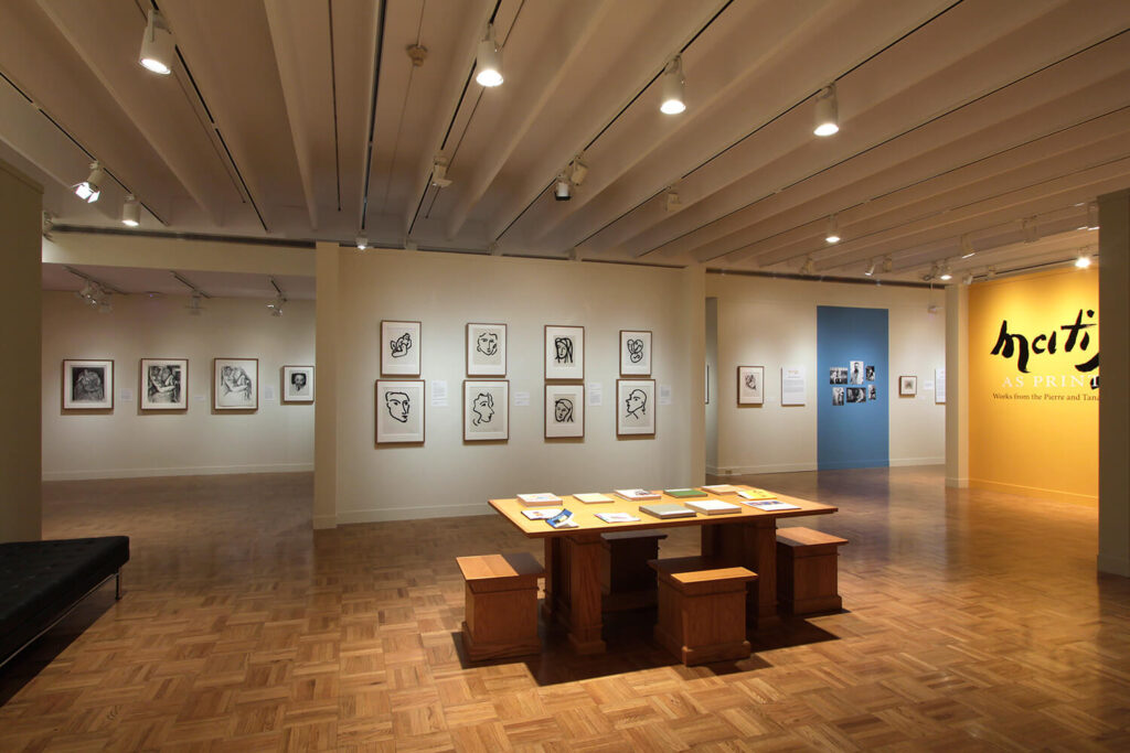 Framed prints by Henri Matisse on walls in a large gallery space. In the foreground is a wooden table with activities on it.