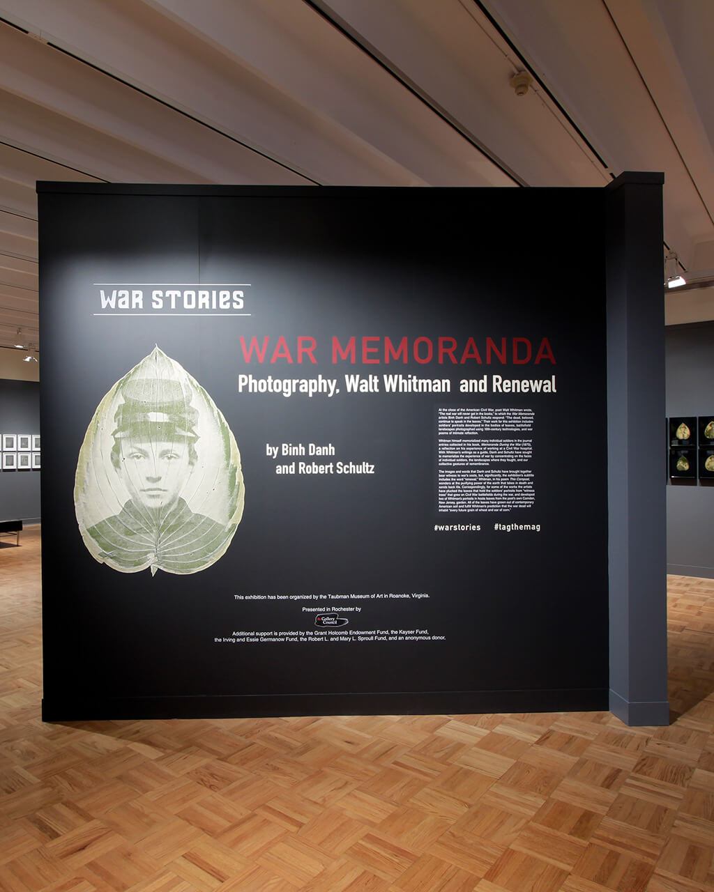 The title wall of the "War Memoranda" exhibition, with text and artwork on a charcoal gray painted wall.