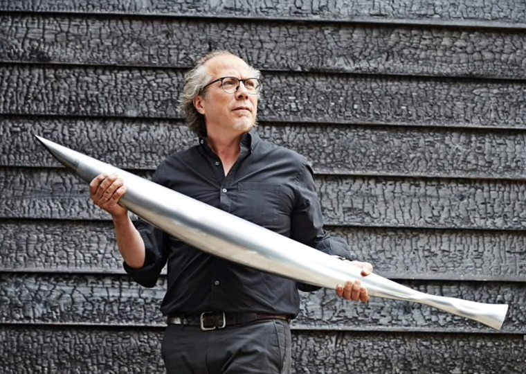 Iñigo Manglano-Ovalle holds a long metal sculpture, looking pensively away from the camera.