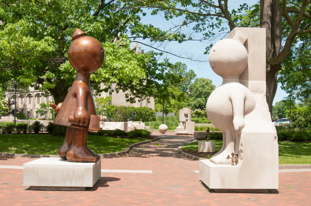 Bronze statue of a stylized female figure with a hammer and chisel looking at a limestone statue of a stylized human still embedded in the block