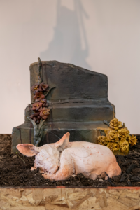 A clay sculpture of a small pig curled up in front of a broken tombstone