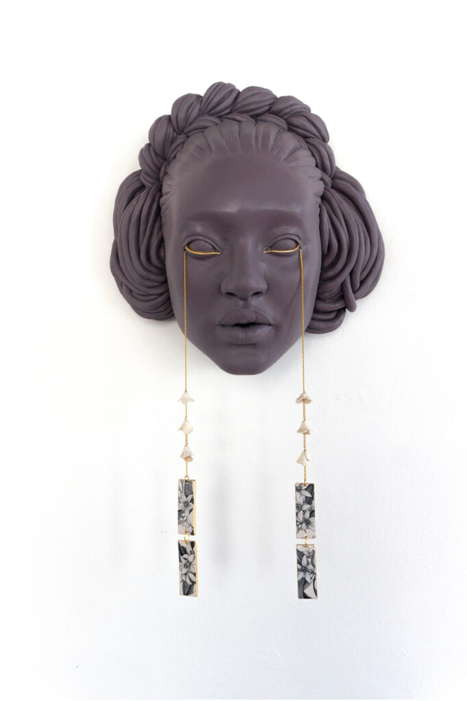 Ceramic sculpture of a woman's face with two gold chains dripping down from the eyes like tears