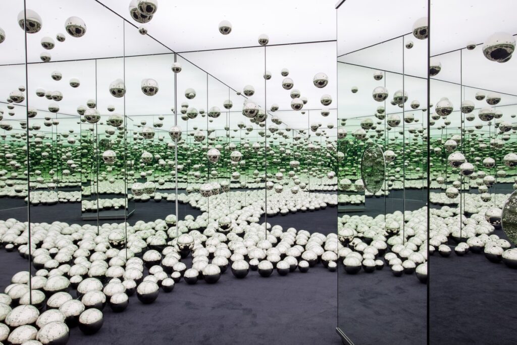 Interior of a mirrored room with a black floor and silver balls.
