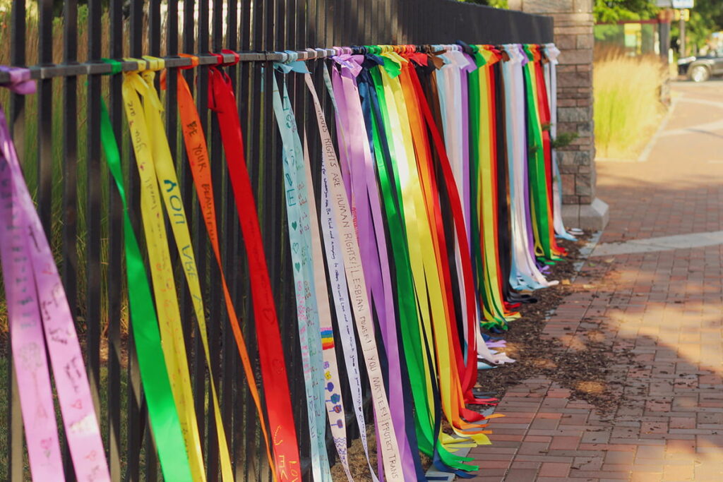 Ribbons with writing on them tied in a rainbow to the fence outside MAG.