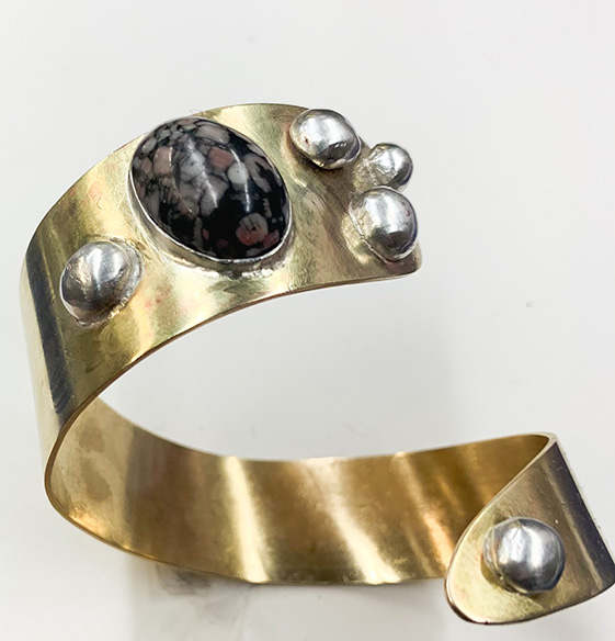 A cuff style bracelet made from brass is twisted into a slight spiral and is ornamented with a single oval shaped black stone and five silver beads at both ends of the cuff.