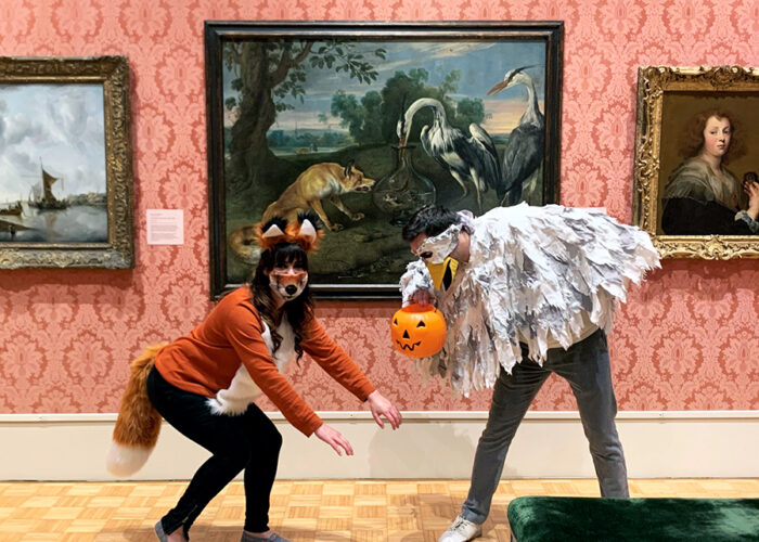 Two people dressed up as a fox and a heron in front of the painting The Fox and the Heron.