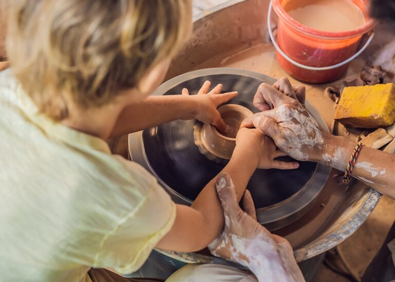 Child throwing a pot on a wheel, with adult hands guiding the student.