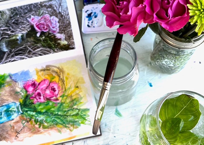 A table displays a series of watercolor palettes, a brush, a vase with pink and green flowers, and a sketchbooks with a painting of the flowers in the vase.