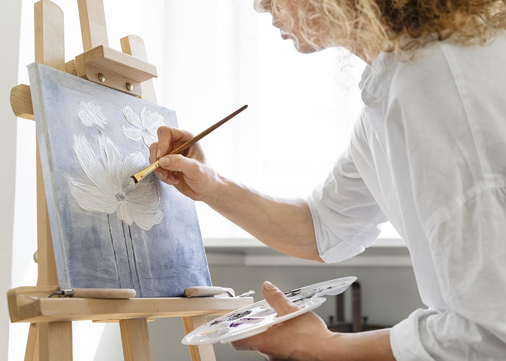 Woman with curly blonde hair painting an image of a white flower on a blue background.