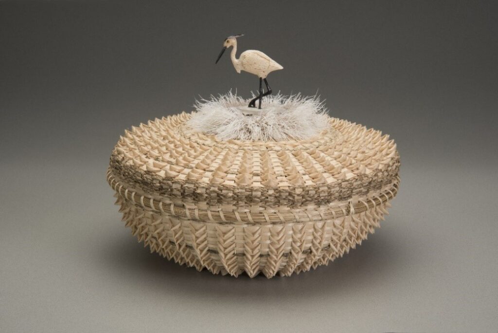 Basket made of black ash wood and braided sweetgrass with a sculpture of a heron on top.