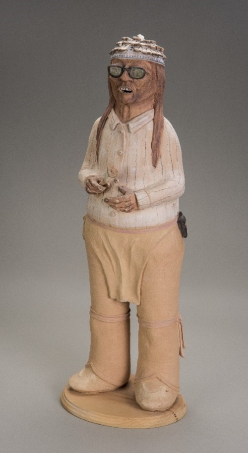 Ceramic sculpture of a man in a traditional Onondaga headdress, holding a horn rattle, a flip phone on his belt.