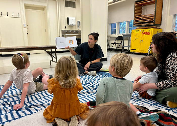 Kids sitting on a blanket in the Creative Workshop, listening to an adult reading a story to them.