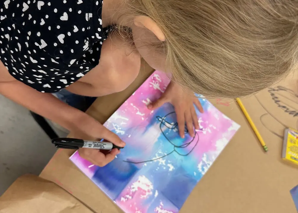 A kid drawing in black marker on paper colored with watercolor.