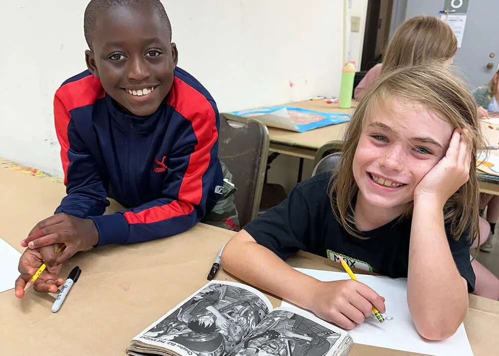 Two kids smiling at the camera while drawing.