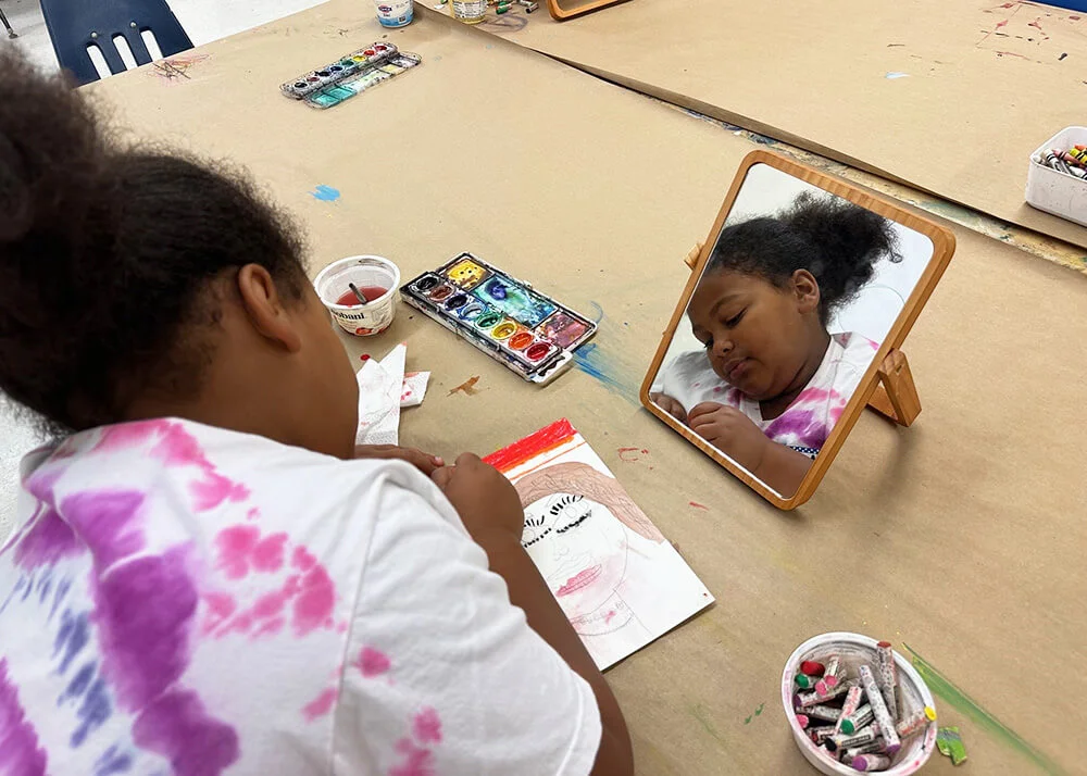 A girl drawing a self-portrait with a table-top mirror.
