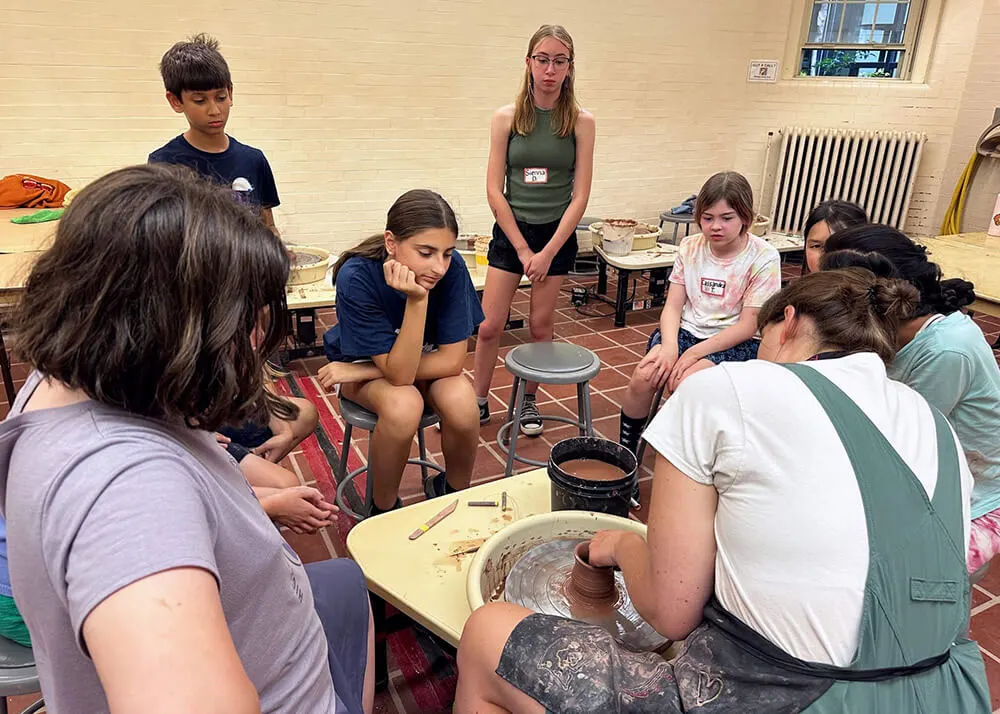 Kids watching a teacher demonstrate throwing a cylinder on a potter's wheel.