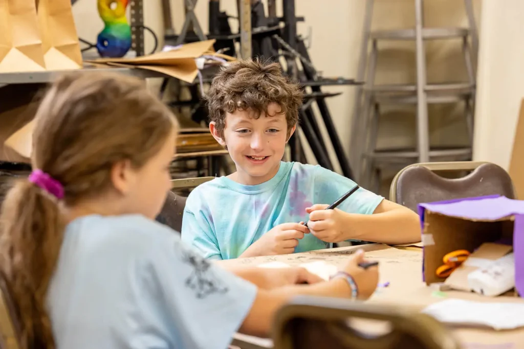 A boy smiling while drawing on a table in the Creative Workshop.