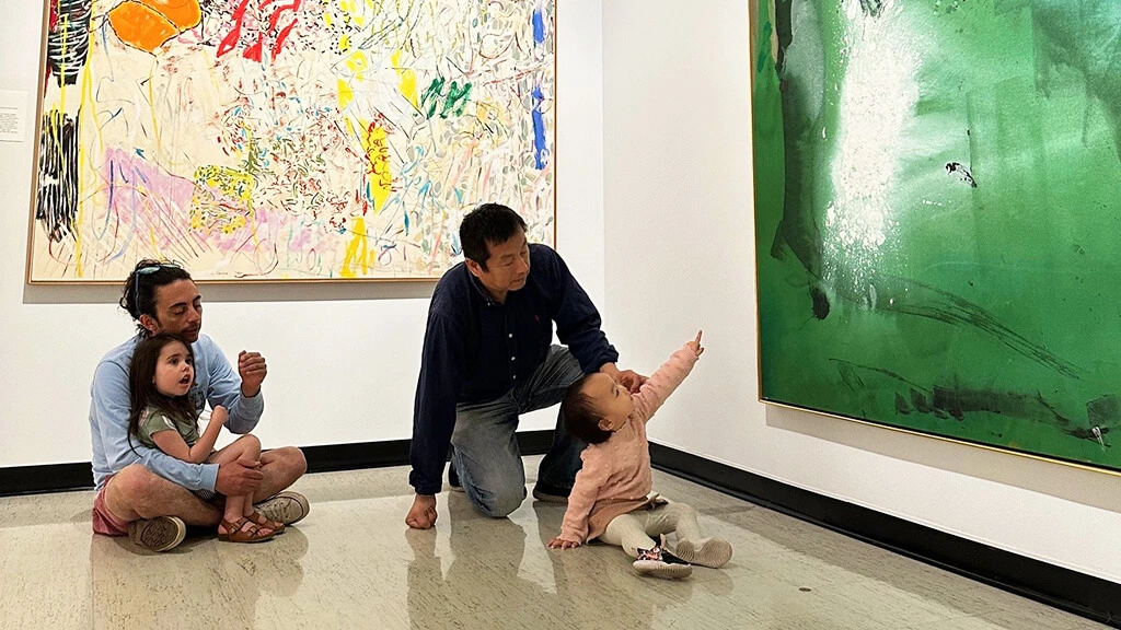Two sets of parents and kids sitting in the galleries, looking at contemporary art paintings.