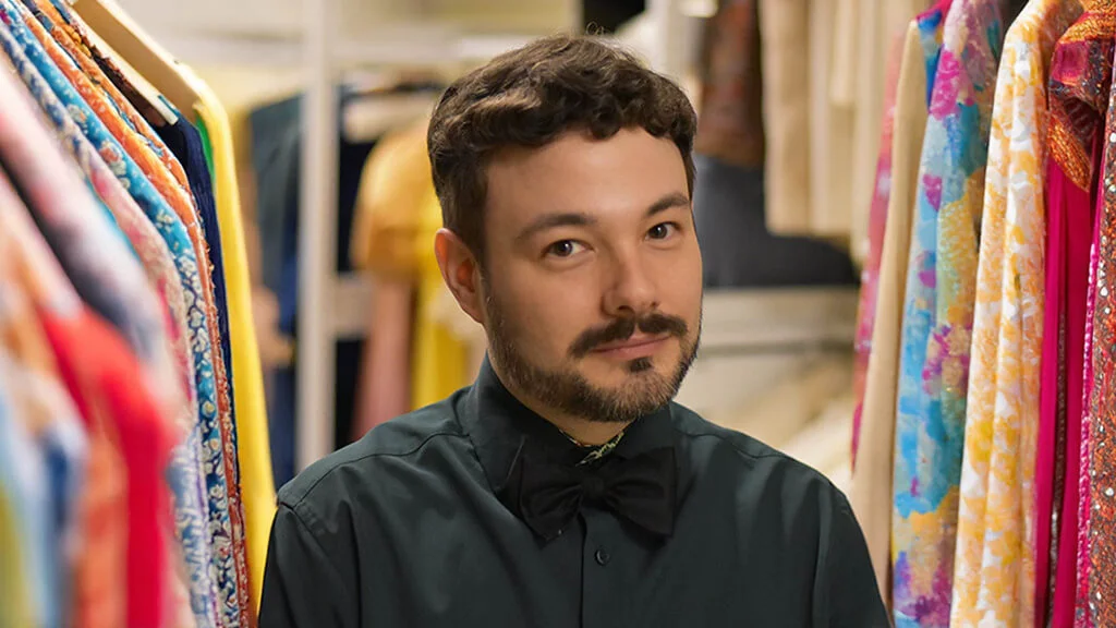 Curator Brandon Brooks with racks of colorful clothing behind him.