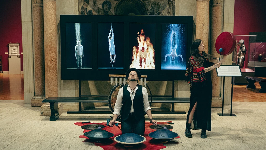 Harold Taddy and Teagan West playing instruments in front of four screens displaying Bill Viola's Martyrs, in MAG's Fountain Court.