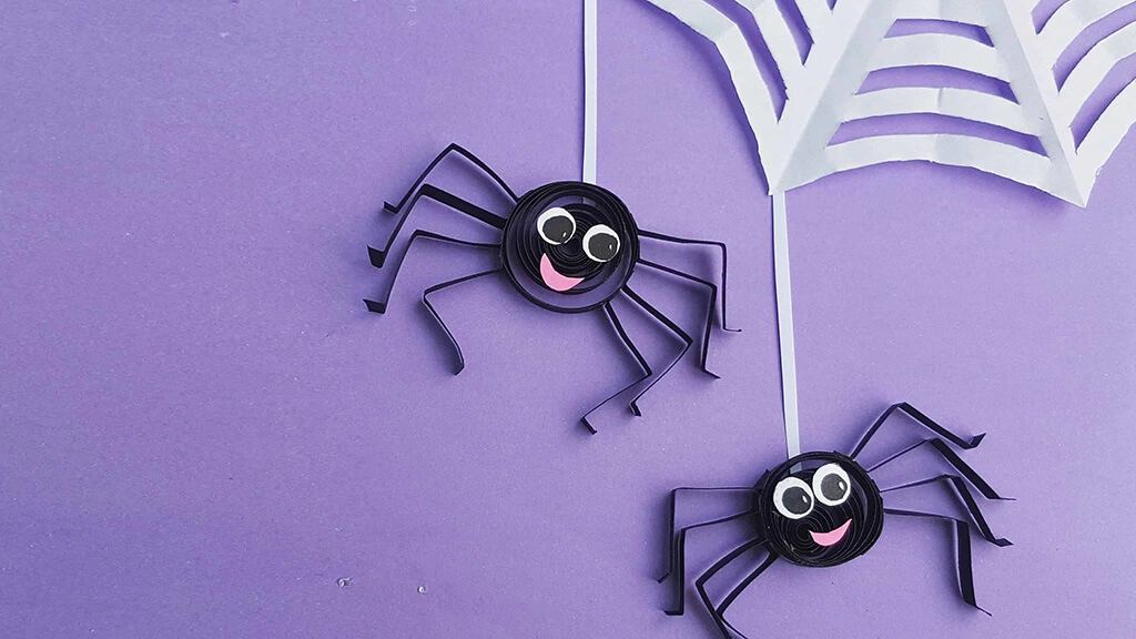 Two black spiders with friendly, cartoon expressions hanging from a white web, all made out of quilled paper.