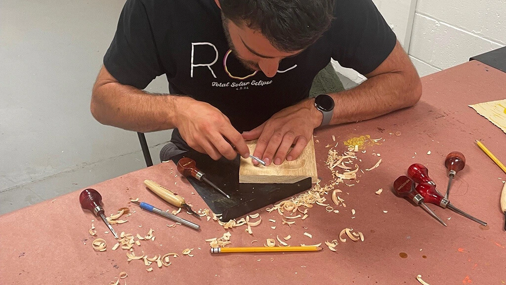 A student works on carving out a woodblock, shavings around his workspace.