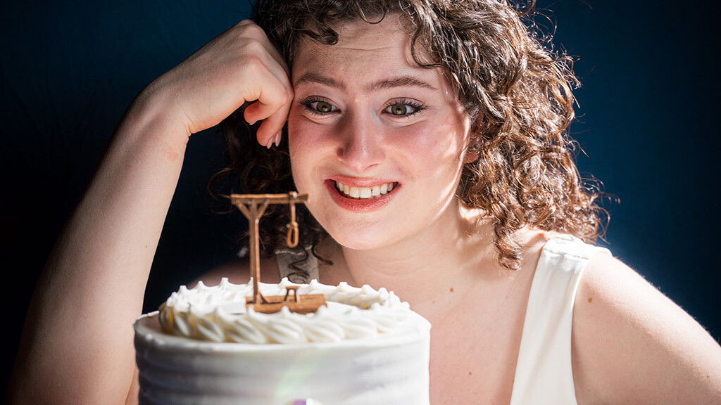 A woman with a forced grin staring at a cake that has a mini gallows topper.