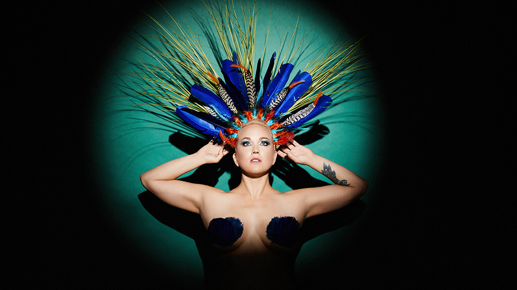 Katie Haverly, haloed in a spotlight and wearing a feathered headdress.
