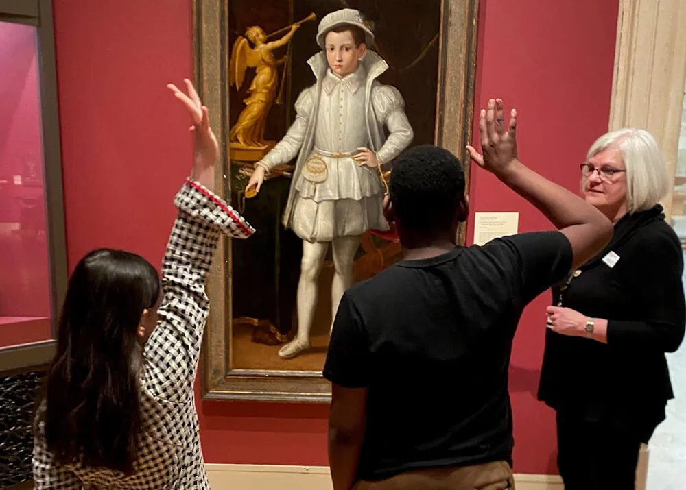 A docent and two children standing in front of a Renaissance portrait painting, the children raising their hands.
