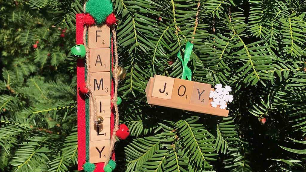 Two Christmas ornaments made out of scrabble tiles, decorated with bells and snowflakes, hanging on pine branches.
