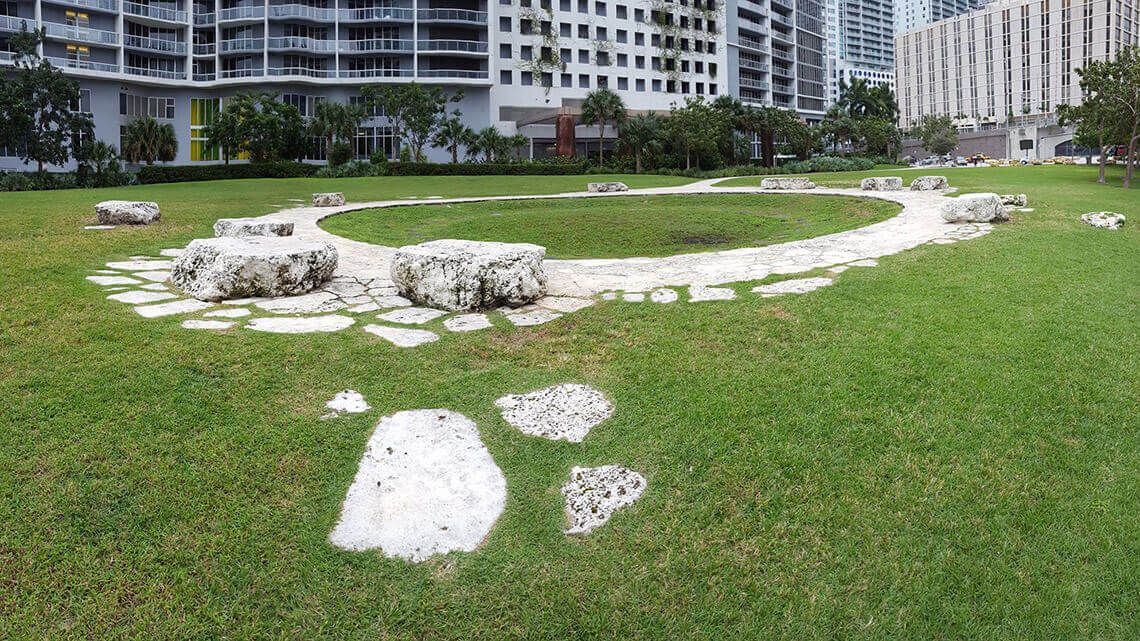 A circle of limestone bedrock revealed in a lawn of green grass, a contemporary office building in the background.