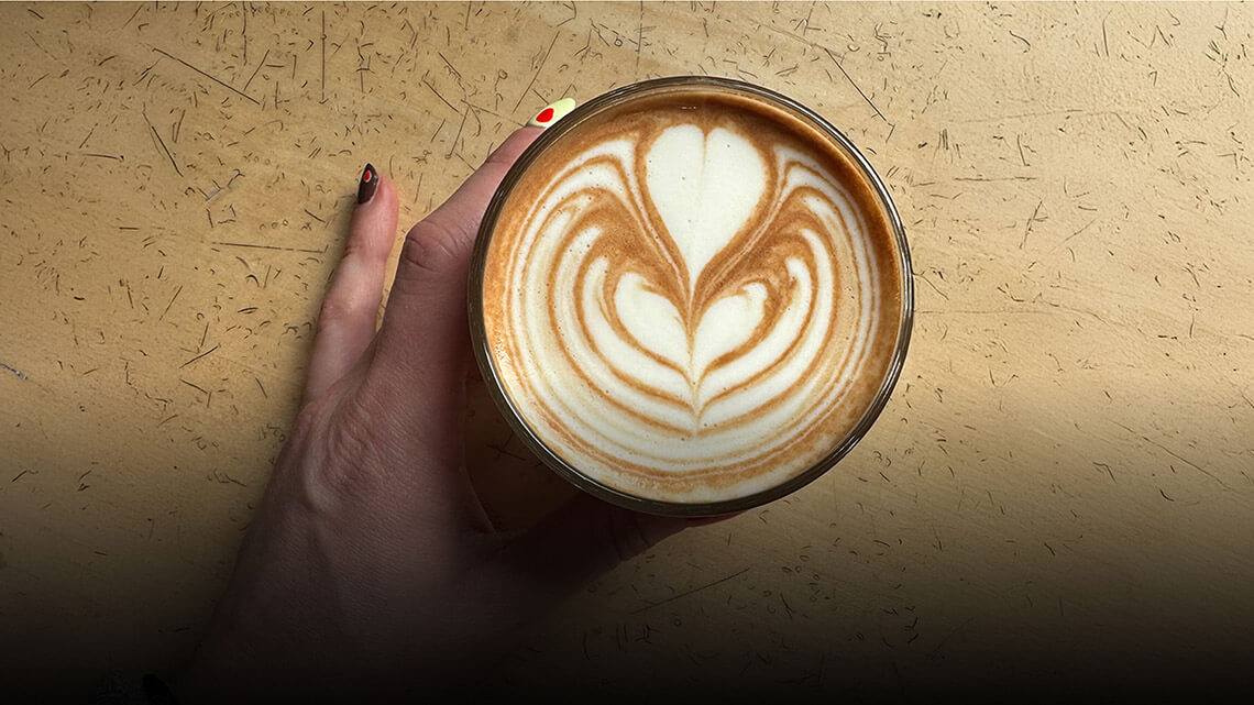A hand holding a cappuccino with heart-shaped latte art drawn in the foam.
