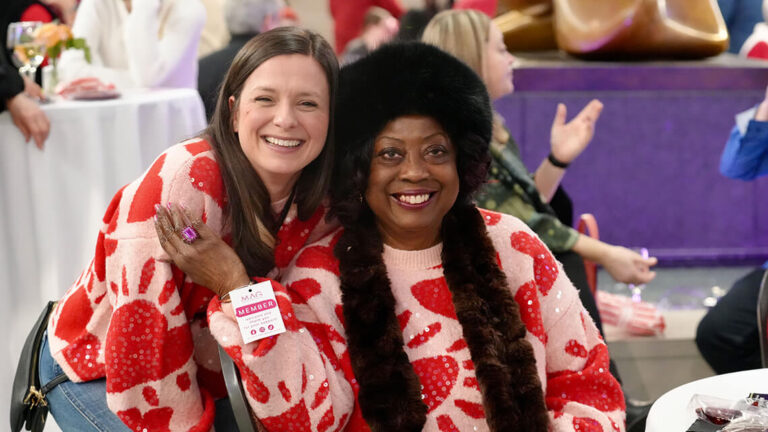 Two women wearing heart sweaters embracing each other and smiling at the camera.