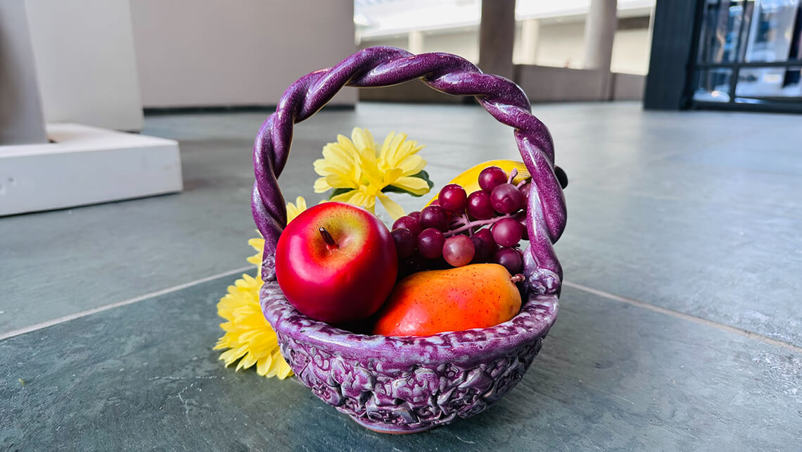 A ceramic basket made to look like it's woven, glazed purple and filled with fruit.
